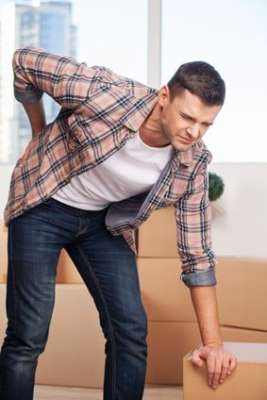 Young man holding hand on his back and expressing negativity while leaning at the cardboard box